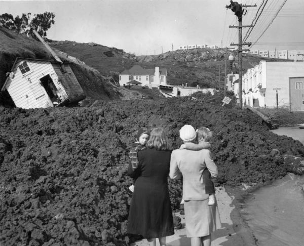 Photographie de deux personnes à côté d’un glissement de terrain ayant recouvert la route et déplacé un bâtiment.