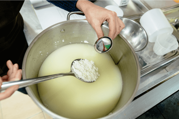 A photograph of a person ladling cottage cheese out of a large saucepan filled with hot milk and vinegar.