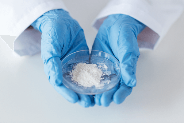 Une poudre blanche dans une boite de Petri dans les mains d’une personne portant des gants bleus et une blouse blanche.